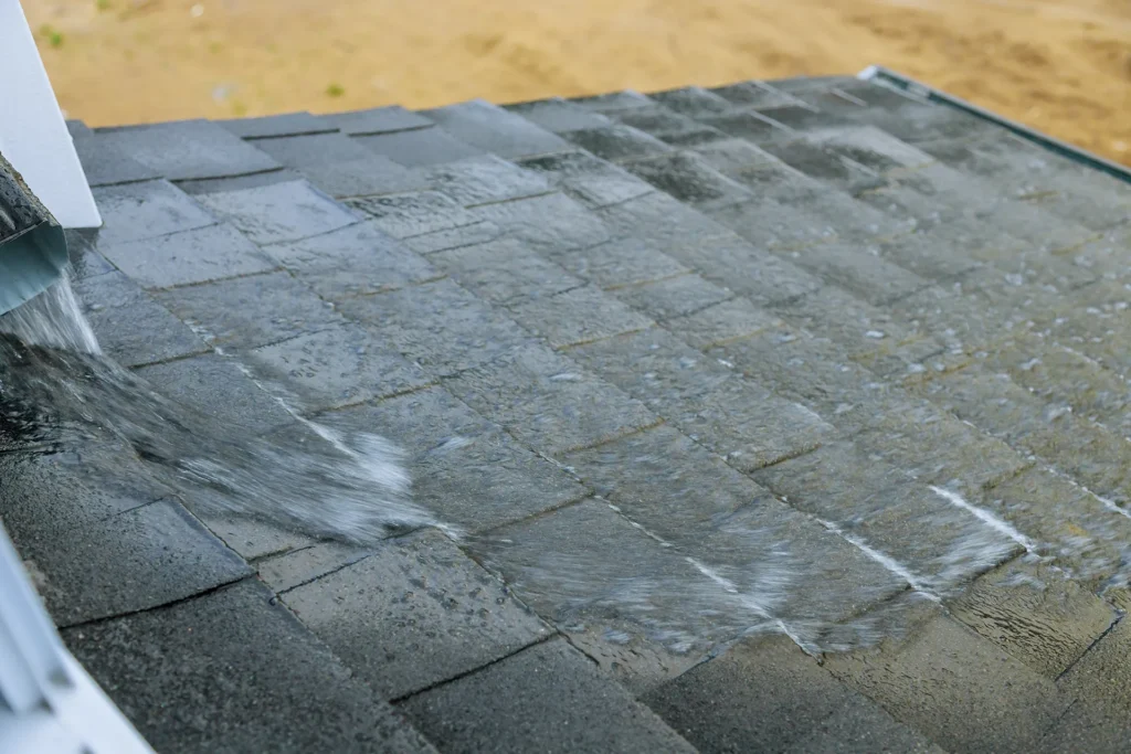 Rainwater flows down a gray asphalt shingle roof, creating a small stream along the surface.