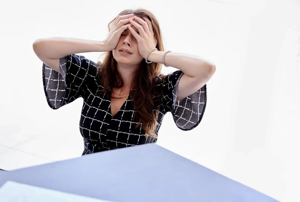 A woman in a black and white patterned dress sits at a table with her hands on her forehead, looking up.