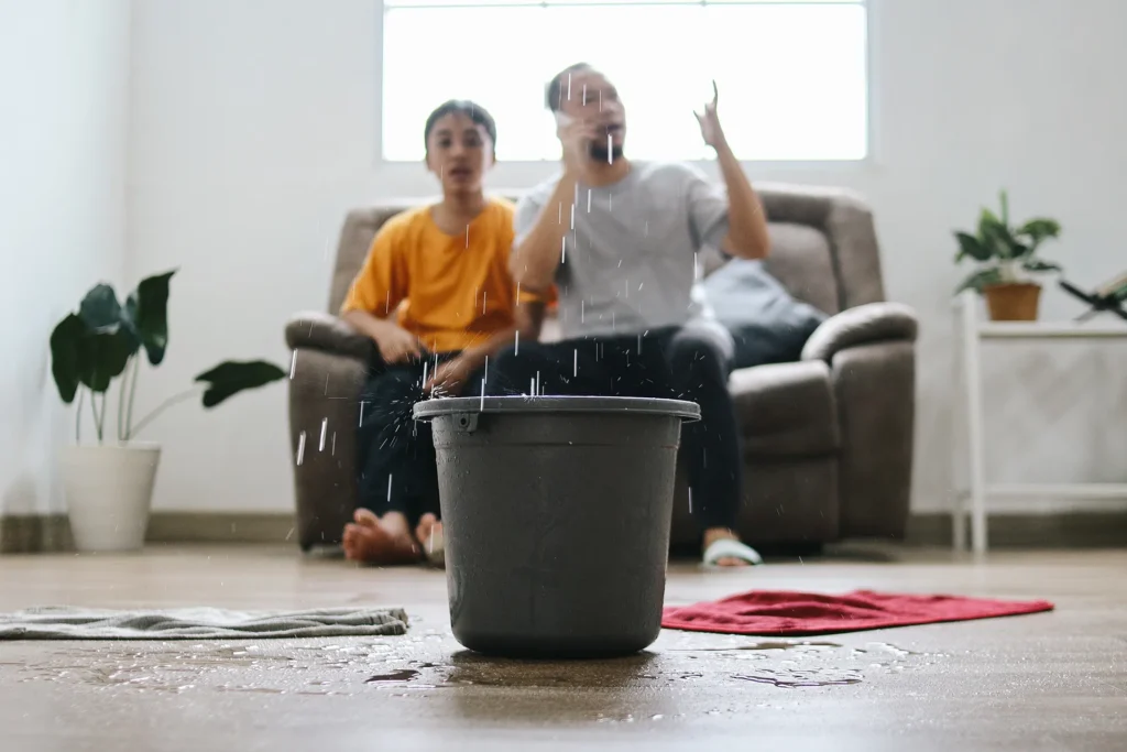 A bucket catches water leaking from the ceiling while two people sit on a sofa in the background, reacting to the leak in a living room.