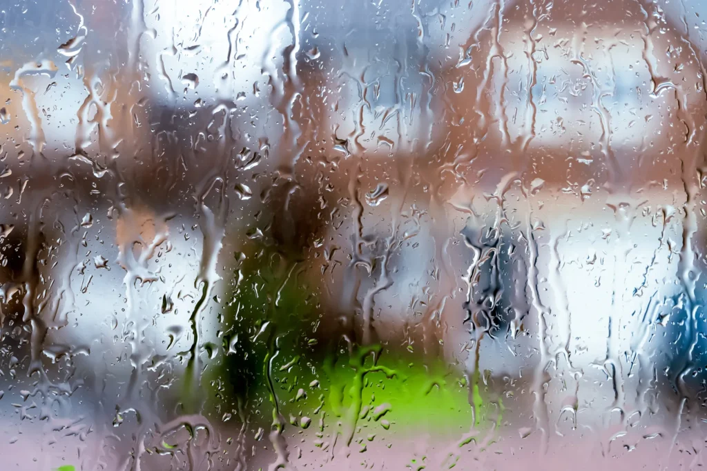 Raindrops on a window with blurred view of houses and greenery outside.